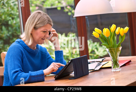 Blonde Frau, die zu Hause im Wintergarten mit ihrem iPad Tablet-Computer und auf ihrem iPhone Smartphone sprechen Stockfoto