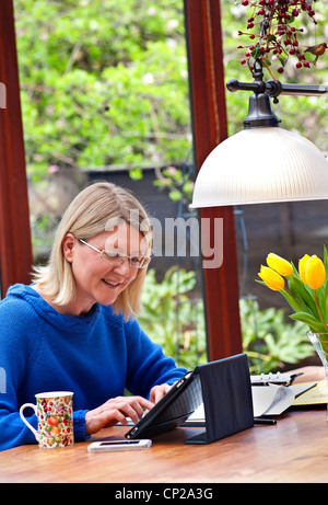 Blonde Frau, die zu Hause im Wintergarten mit ihrem Tablet-Computer iPad in seine Lederbezug Stand arbeiten Stockfoto