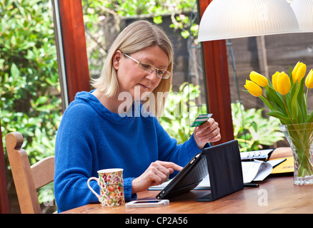 Blonde Frau, die zu Hause im Wintergarten Einkaufen online mit ihrem iPad Tablet-Computer in Leder Case Stand arbeiten Stockfoto