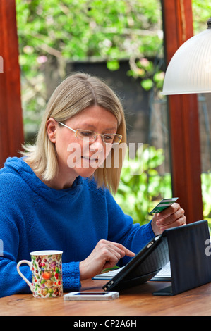 Online-Shopping 40-45 Jahre Blonde Frau zu Hause im Garten Wintergarten Online-Shopping mit ihrem iPad Tablet-Computer in Leder Etui Stand Stockfoto