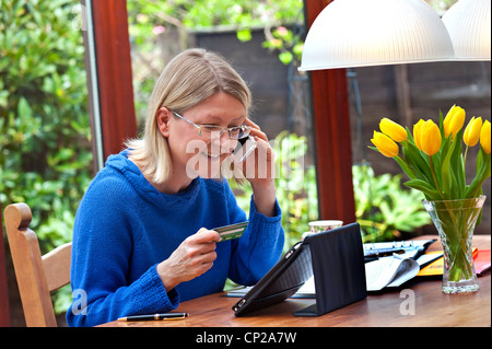 Online-Shopping-Telefon Blonde Frau 40-45 Jahre zu Hause arbeiten Einkaufen online mit ihrem iPad Tablet-Computer und einem iPhone-Smartphone Stockfoto