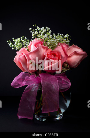 Rosa Rosen mit kleinen weißen Blüten in Glasvase auf dunklem Hintergrund Stockfoto