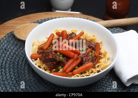 Rindfleisch-Eintopf mit Gemüse über Eiernudeln in White China Schüssel mit Stricken Platzdeckchen & rustikale Holz Löffel im Hintergrund. Stockfoto