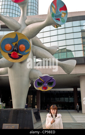 Mädchen auf der Suche auf seinem Mobiltelefon unter eine Skulptur von Farben gebildet von Gesichtern in den Straßen von Tokio, Japan. Stockfoto