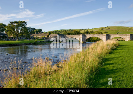 Steinerne Brücke über dem Fluss Wharfe, wie es fließt durch Burnsall Dorf- & malerische Landschaft auf sonnigen Sommerabend - Yorkshire Dales, England, UK. Stockfoto