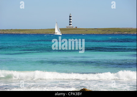 Segelyacht, die im türkisfarbenen Meer am gestreiften Leuchtturm auf Illa de l'Aire vorbeifährt. Vom Strand in Punta Prima, Menorca, Spanien aus gesehen Stockfoto