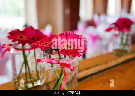 Rosa Gerberas in einer kleinen Vase, ein Glas Konfitüre, gebunden mit rosa Band in einem Spiegel bei einer Hochzeit reflektiert. Stockfoto