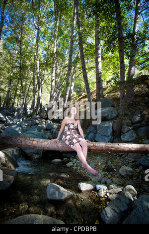 Kaukasische Mädchen mit roten Haaren sitzt auf einem Baumstamm in einem Wald mit einem Lächeln auf ihrem Gesicht. Stockfoto