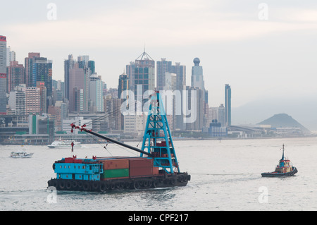 Lastkahn mit Containern geschleppt in den frühen Abendstunden in Hong Kong. Die Skyline von Hong Kong Island im Hintergrund. Stockfoto