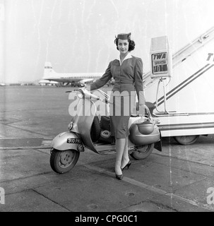 1960er, historisch, eine Werbung aus dieser Zeit, die den neuen Motorroller bewerbt und eine glamouröse TWA-Stewardess an einem Flughafen zeigt, an den Stufen eines Flugzeugs, das mit Helm steht, von ihrem neuen Transportmittel, London Aiport, England, Großbritannien. Stockfoto