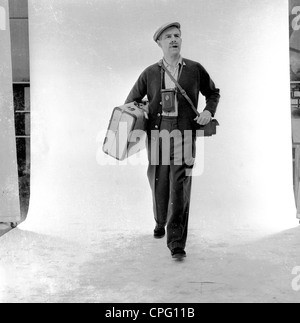 Werbung 1960s. Mann in einem photograhischen Studio, der gegen eine weiße Leinwand steht und mit seinem Koffer, seiner Kamera und seiner Umhängetasche als Urlauber fungiert. Stockfoto