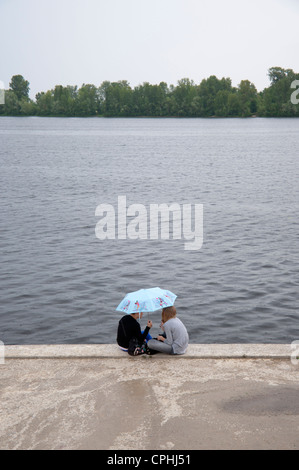 Zwei junge Frauen sitzen unter einem Regenschirm am Fluss Dnjepr, Kiew, Ukraine, Europa. Stockfoto