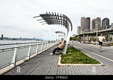 Mann auf Bank mit Schatten Baldachin auf Riverside Park Esplanade verschiebt, als Läufer vor West Side Highway & Skyline von NYC geht Stockfoto