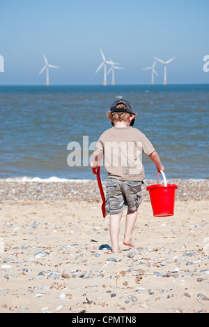 Ein kleiner Junge mit einem Eimer und Schaufel zu Fuß entfernt von der Kamera mit einem Windpark in der Ferne. Stockfoto