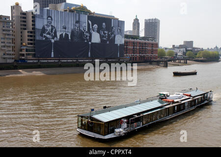 Ein Ausflugsschiff geht von einem riesigen Foto der Königin und der königlichen Familie winken vom Balkon des Buckingham Palace. Das Bild entstand während der 1977 silbernes Jubiläum und ist montiert auf Container Seehaus an der Themse in London als Teil der 2012 Diamant-Jubiläum feiern. Stockfoto