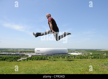 Ein Mann springt auf einem Hügel mit Blick auf die Allianz Arena in München Stockfoto