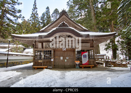 Schneebedeckte schintoistische Schreinhalle aus Holz, mit Eiszapfen, die vom Dachüberhang hängen, und Zedernbäumen dahinter, auf dem berühmten Friedhof von Okunoin in Koya, Japan. Stockfoto