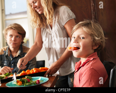 Junge Karotten in den Mund zu halten, als Mutter gesunde Mahlzeit serviert Stockfoto