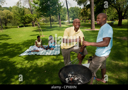 Zwei junge afrikanische Männer Grill zusammen in einem Park mit ihren Freunden sitzen im Hintergrund Stockfoto