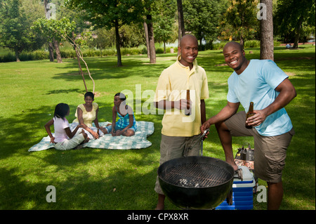 Zwei junge afrikanische Männer Grill zusammen in einem Park mit ihren Freunden sitzen im Hintergrund Stockfoto