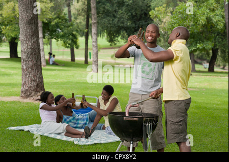 Zwei junge afrikanische Männer Grill zusammen in einem Park mit ihren Freunden sitzen im Hintergrund Stockfoto