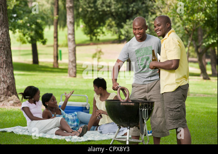 Zwei junge afrikanische Männer Grill zusammen in einem Park mit ihren Freunden sitzen im Hintergrund Stockfoto