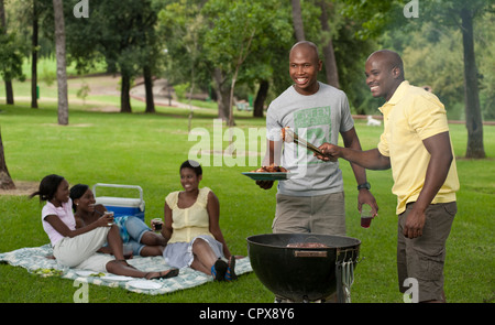 Zwei junge afrikanische Männer Grill zusammen in einem Park mit ihren Freunden sitzen im Hintergrund Stockfoto