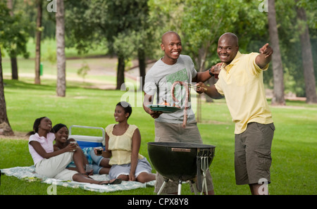 Zwei junge afrikanische Männer Grill zusammen in einem Park mit ihren Freunden sitzen im Hintergrund Stockfoto