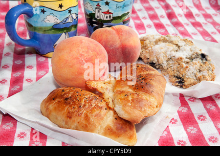 Picknick-Frühstück mit frischen Pfirsichen, Croissants, ein Scone und Kaffee serviert auf einer karierten Tischdecke. Stockfoto