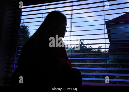 Junge Frau sitzt allein in einem dunklen Raum mit Blick durch ein Fenster Blind. Stockfoto