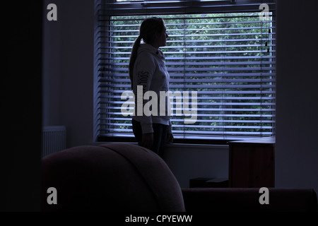 Junge Frau allein in einem dunklen Raum mit Blick durch ein Fenster Blind. Stockfoto