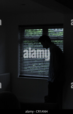 Silhouette eines männlichen stehenden in einem dunklen Raum mit Licht aus einem Fenster mit Jalousien zeigt seine Gestalt. Stockfoto