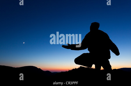 Mann, der auf dem Gipfel eines Berges bei Sonnenuntergang mit dem Mond am Himmel betet. Stockfoto
