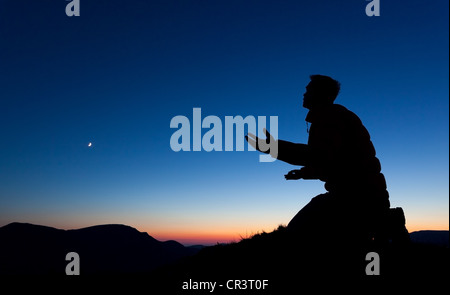 Man bittet Gott auf dem Gipfel eines Berges bei Sonnenuntergang mit dem Mond in den Himmel. Stockfoto