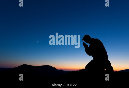 Mann, der auf dem Gipfel eines Berges bei Sonnenuntergang mit dem Mond am Himmel betet. Stockfoto