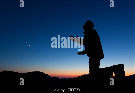 Mann, der betet um Vergebung auf dem Gipfel eines Berges bei Sonnenuntergang mit dem Mond in den Himmel. Stockfoto