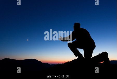 Mann, der betet um Vergebung auf dem Gipfel eines Berges bei Sonnenuntergang mit dem Mond in den Himmel. Stockfoto