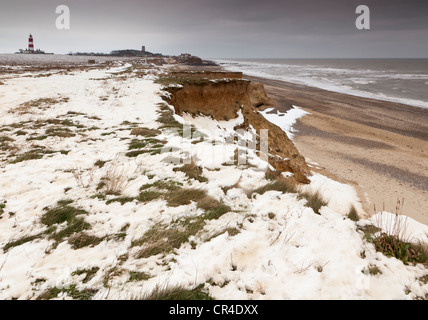 Schnee auf der Klippe und der Strand bei Happisburgh, Norfolk, England, UK, mit Happisburgh Leuchtturm und Kirche in der Ferne Stockfoto