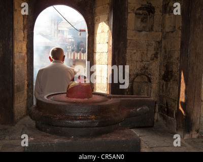 Lokale Mann sitzt hinter einem Shiva Lingam, beobachtete eine Feuerbestattung Zeremonie in Pashupatinath Tempel, Kathmandu, Nepal, Südasien Stockfoto