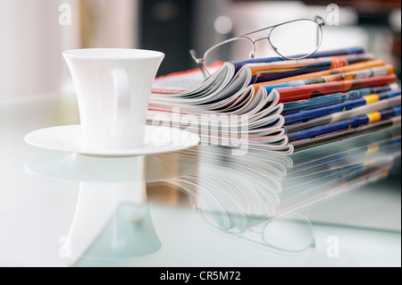 Stillleben mit Zeitschriften, eine Kaffeetasse und ein paar Gläser auf einem Glastisch Stockfoto