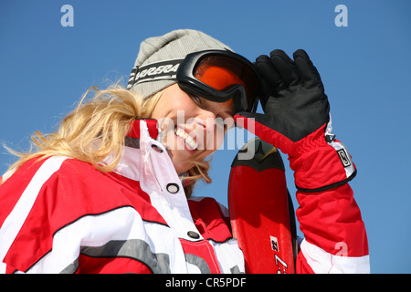 Junge Frau, etwa 25 trägt eine rot-weiße Skianzug und eine blasse graue Mütze gegen einen klaren, blauen Himmel, Lächeln Stockfoto