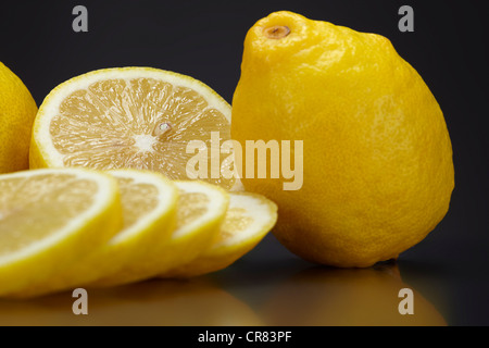 Zitronen (Citrus Limon), ganze Zitrone und in Scheiben geschnittenen Zitronen, auf einer dunklen Glasplatte Stockfoto