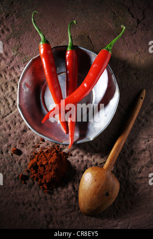 Drei Chili Paprika (Capsicum) auf einem Zinn-Teller mit einem Holzlöffel und Chili-Pulver auf einem rustikalen Steinoberfläche Stockfoto