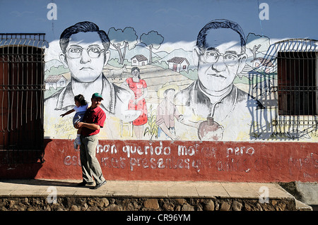 Mann hält ein Kind in seine Arme vor ein Wandbild mit Monsignore Romero und Vater Rafael Palacios, Suchitoto, El Salvador Stockfoto