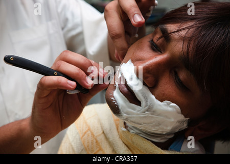 Junge arabische Mann aus Dubai bekommen eine Nassrasur mit einem offenen Rasierer in einem traditionellen Friseure-Shop. Altstadt-Souk. Stockfoto