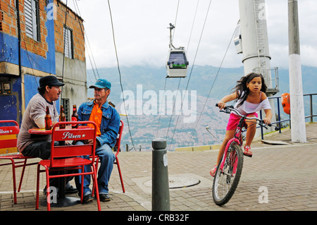 Männer sitzen in einem Straßencafé Mädchen reiten ein Fahrrad, Metrocable Seilbahn am Rücken, Slums, Comuna 13, Medellin, Kolumbien Stockfoto