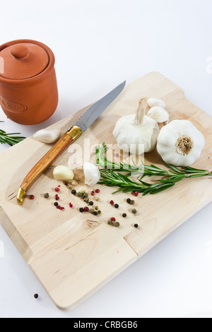 Knoblauch (Allium Sativum) auf einem Holzbrett mit Messer, Römertopf, Rosmarin und schwarzem und rotem Pfeffer Stockfoto