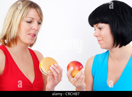 zwei Frauen mit Burger und Apfel. Die dunkelhaarige mit dem Apfel, der Hamburger sowie die blonde Frau mit Interesse betrachten Stockfoto