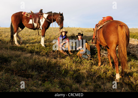 Ein Cowgirl und ein Cowboy sitzt auf dem Boden in der Prärie, ihre Pferde stehen neben ihnen, Saskatchewan, Kanada Stockfoto