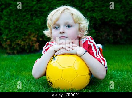 Five-Year-Old Boy ein FC Bayern München Fußball Kleid, mit einem gelben Fußball auf dem Boden liegend Stockfoto